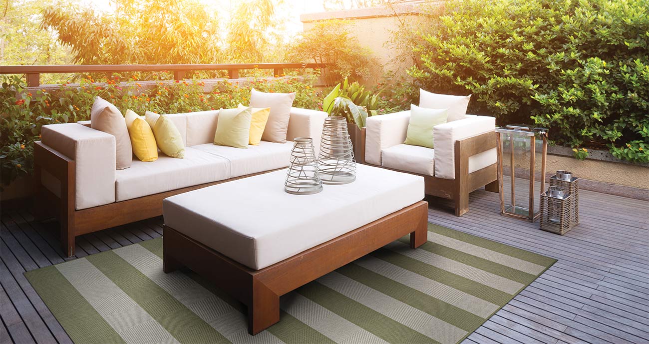 Brown outdoor deck with white couches, sea green and tan area rug, with sunset and bushes in background